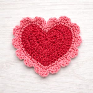 May include: A handmade, crocheted heart-shaped coaster. The center is a deep red, surrounded by a lighter pink scalloped edge. The coaster is made of yarn and sits on a white wooden surface.