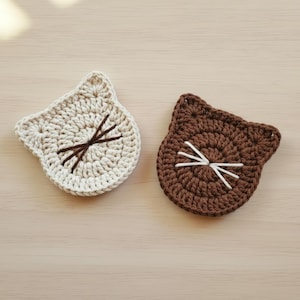 May include: Two handmade crochet cat-shaped coasters. One is off-white with brown embroidered whiskers, and the other is brown with white whiskers. The coasters are round with pointed ears, perfect for a cat lover's home decor.