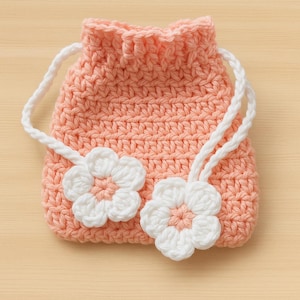 May include: A peach-coloured crochet drawstring bag with two white flower appliqués. The bag has a white drawstring closure and is made of a textured yarn. The flowers have a peach centre and white petals.