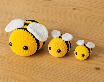 Può includere: Tre peluche a forma di ape all'uncinetto di diverse dimensioni. Le api sono gialle con strisce nere e ali bianche. Ogni ape ha occhi neri a bottone. Sono disposte su una superficie di legno marrone chiaro.