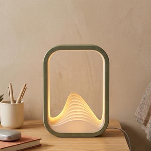 May include: A modern, olive green framed lamp with a layered, wave-like design that emits a warm glow. The lamp sits on a wooden surface next to a small potted succulent, pens, and a book. A silver glasses case rests on the book.