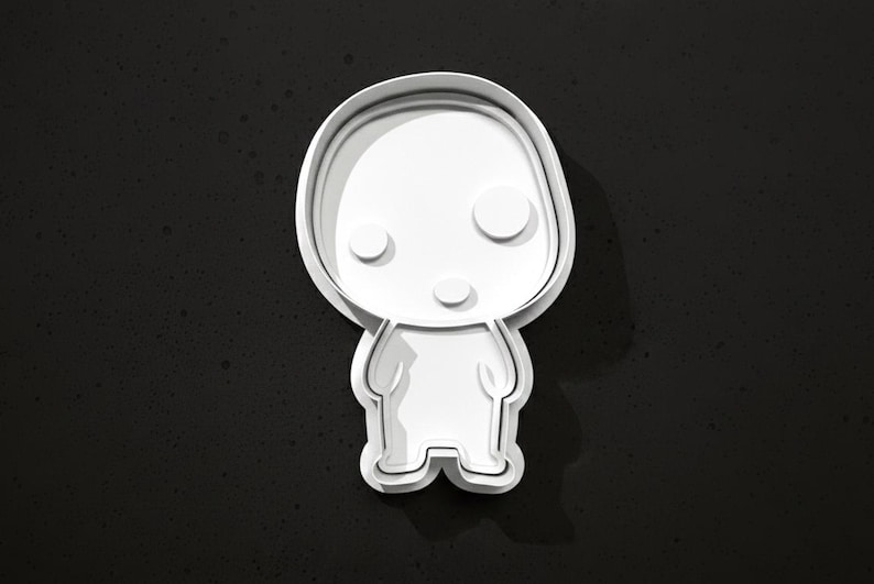 May include: A white, layered cookie cutter in the shape of a stylized cartoon character with a rounded head, two circular eyes, and a small mouth. The cutter is set against a dark, speckled background.