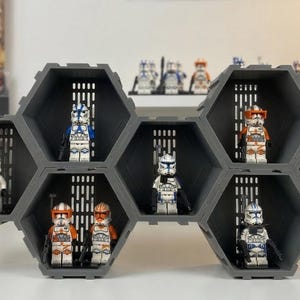 May include: A gray, hexagonal display shelf holding various Star Wars Lego figures. The shelf is designed with multiple compartments, each showcasing a different Lego character. The figures are primarily clone troopers in white, blue, and orange armor.
