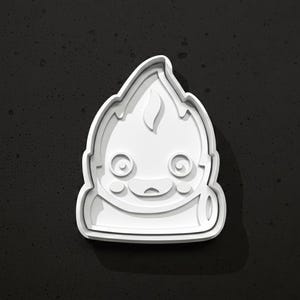May include: A white plastic cookie cutter in the shape of a cartoon character with a flame-like head. The cutter has a smiling face with large eyes and a small mouth. The design is on a dark gray background.