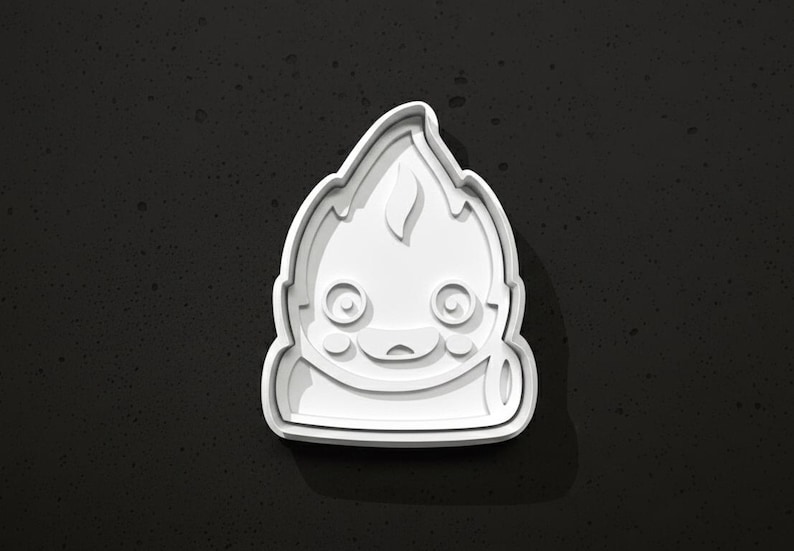 May include: A white plastic cookie cutter in the shape of a cartoon character with a flame-like head. The cutter has a smiling face with large eyes and a small mouth. The design is on a dark gray background.