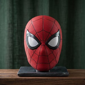 May include: A red Spider-Man mask featuring a textured surface with a black web pattern and large, white, mesh-covered eye lenses framed in black. The mask is mounted on a square, dark gray base, displaying a detailed, comic-inspired design.