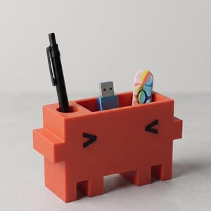 Pode incluir: Um organizador de secretária retangular e sólido na cor coral, com dois olhos pretos em forma de chevron. Possui um orifício circular que contém uma caneta preta e uma ranhura retangular com uma pen drive azul e um pequeno objeto oval multicolorido.