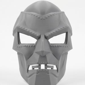 May include: A gray, rigid, full-face mask featuring a stylized, angular design with prominent geometric contours, a sharp nose bridge, wide eye openings, and a distinct mouth section with visible rivet-like detailing along the brow and jawline.