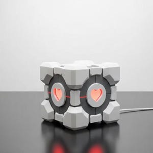Könnte beinhalten: Eine grau-schwarze, würfelförmige Lampe mit einem Herzdesign auf der Vorderseite. Die Lampe hat einen roten Schein, der von den Herzformen ausgeht. Die Lampe ist an eine Steckdose angeschlossen.