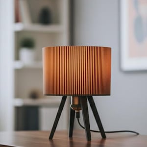 May include: A modern table lamp with a cylindrical, ribbed shade in warm orange tones. The lamp sits on a tripod base with black legs and a brass-colored central support. The lamp is plugged into an outlet.