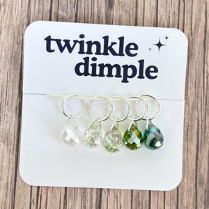 Pode incluir: Um cartão branco com o texto "twinkle dimple" e cinco anéis de prata, cada um segurando um pingente de cristal facetado em tons claros, iridescentes e verdes. Os pingentes têm formato de gota.