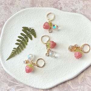 May include: Four gold-tone hoop earrings, each featuring a small pink strawberry charm, a clear faceted crystal bead, and a tiny clear rhinestone accent. The charms are attached to the hoops with small gold-tone jump rings and metal findings.