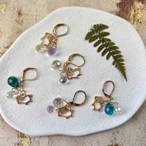 Op de afbeelding: Vijf paar goudkleurige oorringen met hangers van iriserende en gekleurde kristallen kralen. Elke oorring heeft een kleine, opengewerkte varkensbedel. De oorbellen worden gepresenteerd op een wit, organisch gevormd oppervlak met een varenblad.