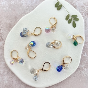 Può includere: Una collezione di orecchini a leva color oro, ciascuno ornato con grappoli di perline di cristallo colorate e sfaccettate. Gli orecchini presentano una varietà di colori, tra cui blu, rosa, trasparente e verde, su una superficie bianca di forma organica.