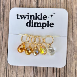 Puede incluir: Una tarjeta blanca con el texto "twinkle dimple" y un conjunto de seis marcadores de punto dorados. Cada marcador tiene una cuenta de cristal facetada en forma de lágrima en tonos naranja, amarillo y transparente. Los marcadores están unidos a un hilo blanco.