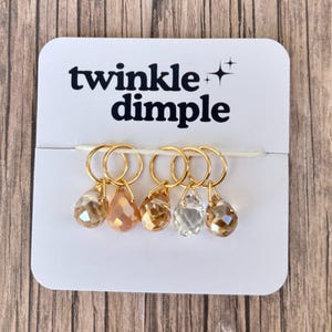 Puede incluir: Un conjunto de cinco marcadores de punto dorados con colgantes de cristal en forma de lágrima en tonos melocotón, ámbar y transparente. Los marcadores están sujetos a una tarjeta blanca con el texto "twinkle dimple".