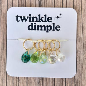 Puede incluir: Una tarjeta blanca con el texto "twinkle dimple" en letras negras. Se adjuntan seis anillos dorados, cada uno con una cuenta facetada en forma de lágrima en tonos verdes y claros. La tarjeta está sobre una superficie de madera.