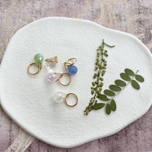 May include: A collection of gold-toned rings, each featuring a different colored gemstone. The gemstones include green, clear, blue, pink, and iridescent white. The rings are arranged on a white, textured surface, with green foliage accents.