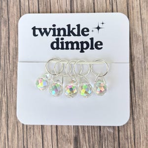 Op de afbeelding: Een witte kaart met de tekst "twinkle dimple" en een sterrenafbeelding bevat vijf heldere, facetgeslepen kristallen bedels met zilverkleurige ringen. De bedels hebben een regenboog iriserend effect. De achtergrond is een houten oppervlak.