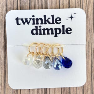 May include: A set of five gold stitch markers with teardrop-shaped, faceted crystal beads in white, clear, grey, blue, and navy blue. The card reads "twinkle dimple" in black text.