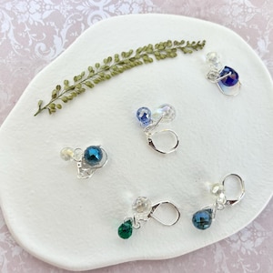 May include: An assortment of dangle earrings with silver-tone hooks and various colored crystal beads. The earrings feature clear, blue, and green faceted beads. A sprig of dried greenery rests on a white, organic-shaped surface.