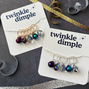 May include: Two white display cards labeled twinkle dimple, each holding five teardrop-shaped faceted crystal charms attached to metal rings. One card features gold-toned rings, while the other displays silver-toned rings. The crystals are purple, iridescent, clear, blue, and silver.
