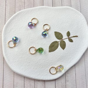 Puede incluir: Varios pendientes de aro dorados con cuentas de cristal facetadas e iridiscentes de varios colores se disponen sobre una superficie blanca texturizada. Una hoja de rosa seca añade un elemento natural. Los pendientes son para orejas perforadas.