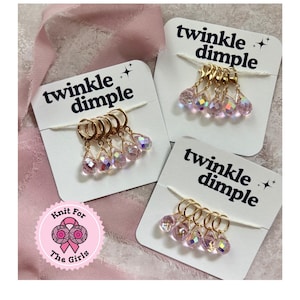 Puede incluir: Tres juegos de marcadores de punto dorados con dijes de cristal rosa iridiscente en forma de lágrima. Cada juego está unido a una tarjeta blanca con el texto "twinkle dimple". También es visible un logotipo rosa con el texto "Knit For The Girls".