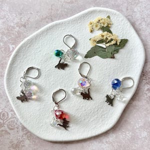 Puede incluir: Un conjunto de pendientes de plata con cierre de palanca, cada uno con un pequeño dije de silueta de gato, cuentas iridiscentes y un cristal de colores. Los pendientes se exhiben sobre un plato blanco texturizado con flores secas.