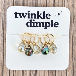 May include: A white card with the text "twinkle dimple" and five gold-colored hoop earrings, each with a faceted, teardrop-shaped crystal pendant. The crystals are clear, smoky, and iridescent. The earrings are attached to the card with a white string.