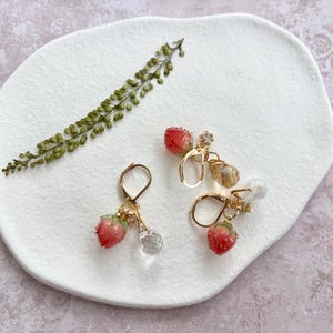 Könnte beinhalten: Zwei Paar goldfarbene Ohrhänger. Jeder Ohrring hat einen kleinen, roten Erdbeeranhänger mit einer klaren, facettierten Perle und einem kleinen, klaren Kristall. Ein Zweig grünes Laub ist ebenfalls vorhanden.