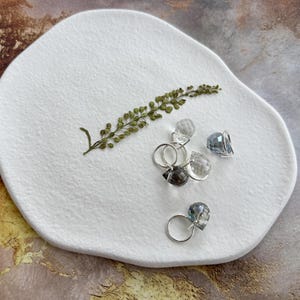Puede incluir: Un conjunto de anillos de piedras preciosas con bandas de plata, exhibidos sobre una placa blanca de forma orgánica. Los anillos presentan piedras de cuarzo facetadas, claras y ahumadas. Una ramita de vegetación seca añade un acento natural.
