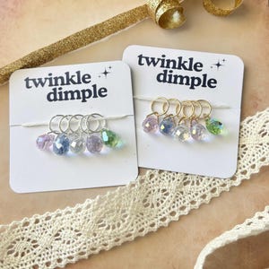 May include: Two sets of stitch markers on white cards with the text "twinkle dimple". Each set has five teardrop-shaped beads in pastel colors. One set has silver rings, the other has gold rings. A gold ribbon and lace trim are in the background.