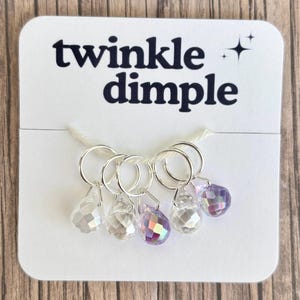 Puede incluir: Una tarjeta blanca con el texto "twinkle dimple" y una estrella. Se adjuntan cinco anillos plateados, cada uno con un dije de lágrima de cristal facetado. Los dijes son transparentes y de color púrpura iridiscente.