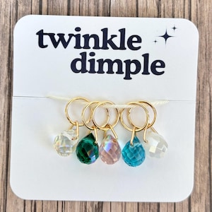Puede incluir: Una tarjeta blanca con el texto "twinkle dimple" presenta seis pendientes de aro dorados, cada uno con un colgante de cristal en forma de lágrima de diferente color: transparente, verde, rosa, turquesa y blanco. La tarjeta está sobre una superficie de madera.