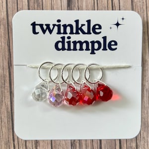 May include: A white card with the text "twinkle dimple" and a star graphic. Attached are five silver stitch markers with teardrop-shaped beads in clear, iridescent pink, and red. The markers are strung on a white thread.