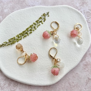 Puede incluir: Cuatro pares de pendientes dorados con dijes de fresa. Las fresas son rosas con hojas verdes y detalles dorados. Cada pendiente tiene una cuenta transparente facetada y un cierre dorado. Los pendientes se exhiben sobre una superficie blanca texturizada.