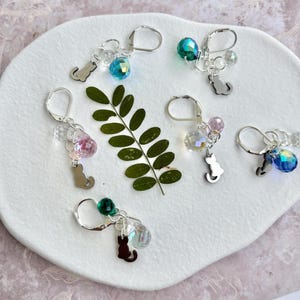 May include: A collection of silver leverback earrings with cat charms and colorful crystal beads. The earrings feature various colors, including blue, pink, and green, and are arranged on a white, textured surface with a green leaf.
