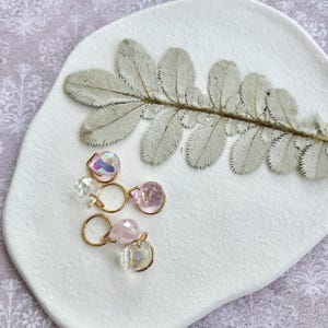 Puede incluir: Pendientes dorados con colgantes de cristal iridiscente y rosa. Se muestran sobre un plato blanco texturizado, con una hoja de helecho seca. El fondo es una tela morada clara con un estampado floral.