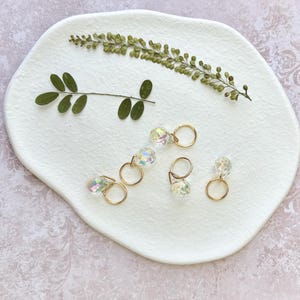 Puede incluir: Pendientes de aro dorados con colgantes de cristal iridiscente, presentados sobre una placa de cerámica blanca de forma orgánica. Follaje verde seco acentúa los pendientes, creando una presentación natural y elegante. El diseño es minimalista.