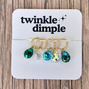 Puede incluir: Una tarjeta blanca con el texto "twinkle dimple" muestra seis anillos dorados, cada uno con una cuenta facetada en forma de lágrima. Las cuentas son de varios tonos de verde, transparente e iridiscente.
