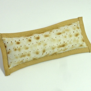 Op de afbeelding: Een rechthoekig kussen met een beige rand en een patroon in het midden dat lijkt op matzah. De matzah-achtige stof heeft een lichtbeige achtergrond met bruine vlekken en perforaties, waardoor het een textuur krijgt. Het kussen is een decoratief item.