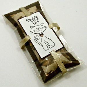 May include: A clear cellophane package containing a rectangular item with a brown border and a tan patterned center. A white tag with a cat illustration and the text "thedots.etsy.com" is attached with a gold ribbon.