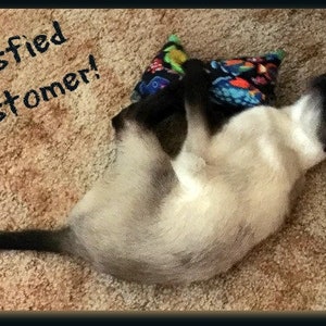 May include: A Siamese cat with cream-colored fur and dark points is lying on its back, paws up, next to a patterned pillow. The text "Satisfied Customer!" is written above the cat.