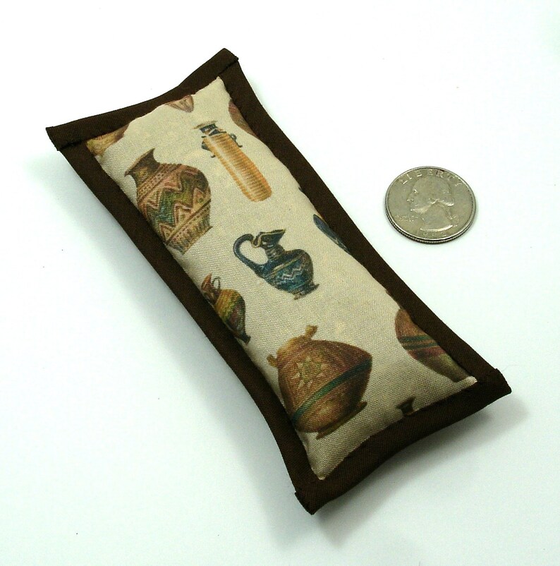 May include: A rectangular cat toy with a brown border. The beige fabric is printed with colorful illustrations of ancient pottery. A quarter is next to the toy for size comparison.