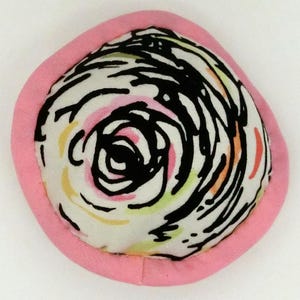 May include: A pink fabric covered round pincushion with a white fabric center featuring a black and white swirl design with hints of pink, orange, and green.