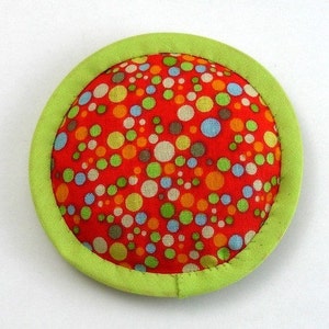 May include: A round, red fabric toy with a green border. The fabric has a polka dot pattern with various colours, including red, orange, yellow, green, blue, and white.