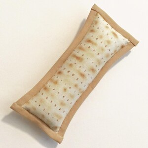 Op de afbeelding: Een rechthoekig, beige stoffen item met een patroon van bruine stippen, dat op matzah lijkt. De randen zijn afgewerkt met een lichtbruine stof. Het item is diagonaal geplaatst tegen een witte achtergrond.