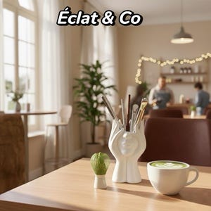 Peut inclure: Un porte-ustensiles en céramique blanche avec un visage, contenant divers ustensiles. Un fouet à matcha et une tasse de matcha latte sont sur une table en bois. Le texte "Éclat & Co" est en haut.