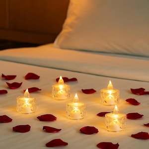 May include: A romantic scene featuring several battery-operated LED candles with a flickering flame effect. The candles are clear and sit on a white surface scattered with red rose petals. The background includes a white pillow and bedsheet.